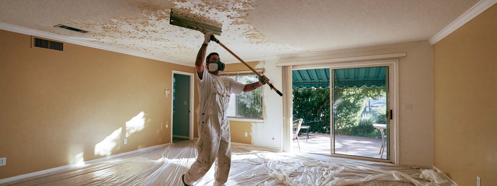 A contractor scraping a sprayed ceiling in a cleared room, plastic sheeting across the whole floor