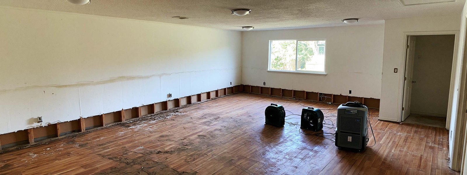 An emptied room after a leak, the bottom two feet of wall cut away to expose the studs, two air movers and a dehumidifier running