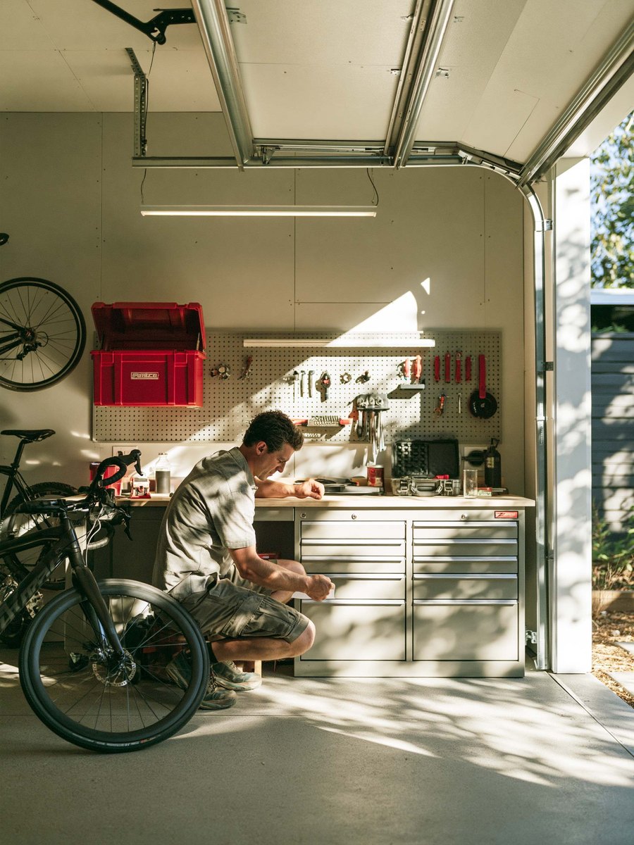 A man crouched by a workbench in a bright finished garage, bicycles beside him and the door open