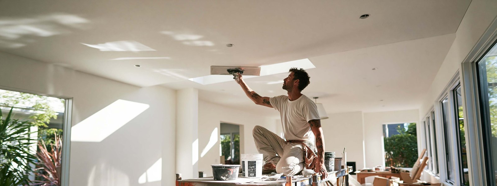 A drywaller kneeling on a plank, smoothing a ceiling with a trowel in a bright room