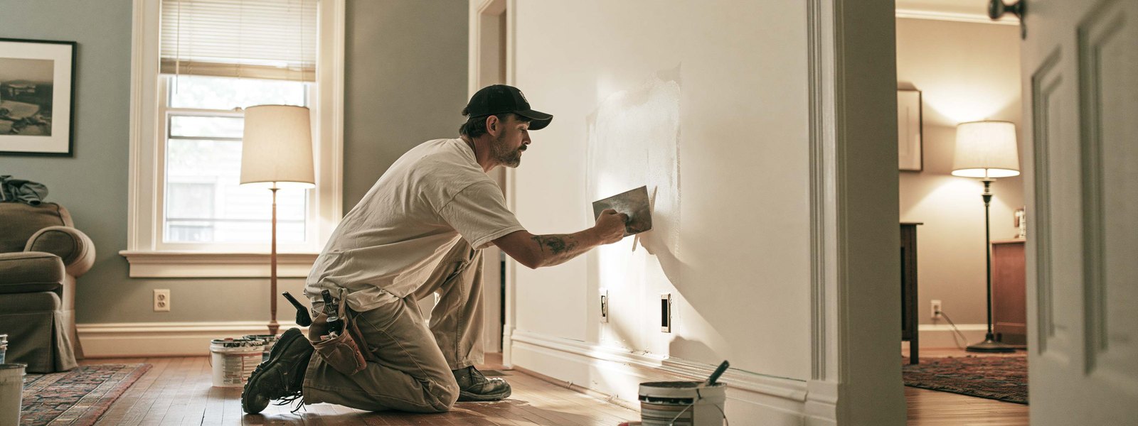 A drywaller kneeling to smooth a repaired area on a living room wall, lamps lit in the rooms beyond